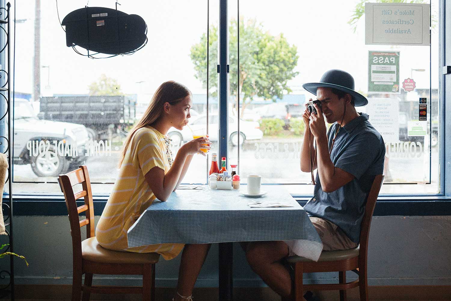 woman sitting at table drinking through a straw and man sitting across table taking her photo with a camera