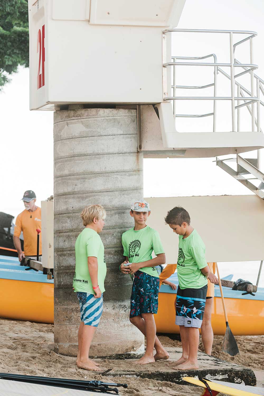 Three little boys in green shirts and board shorts stand by a life guard station
