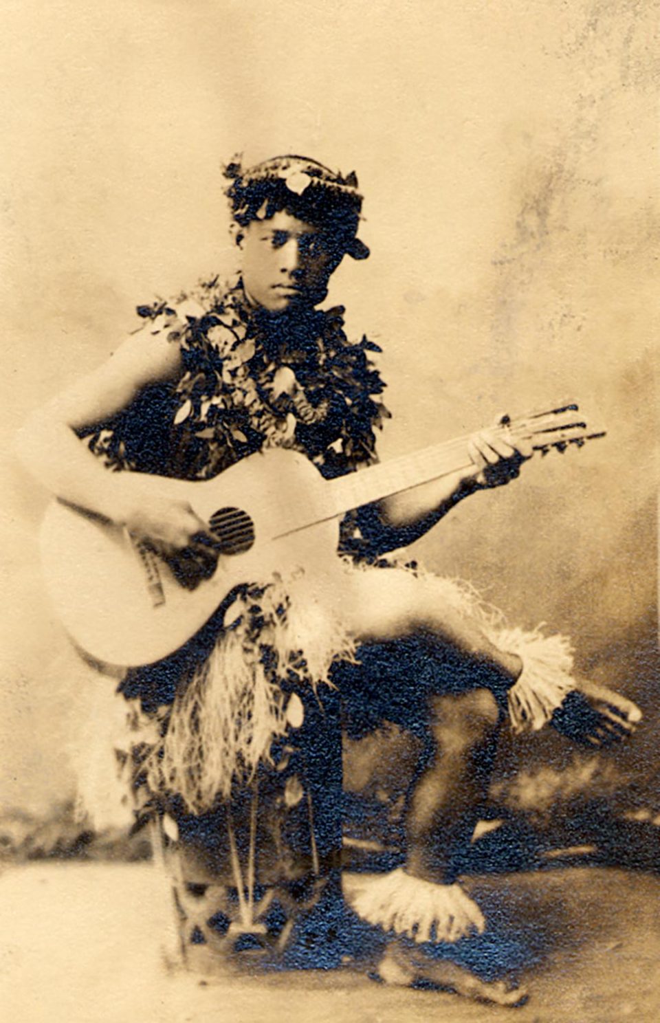 Photo of a young man dressed with various lei and holding a guitar.